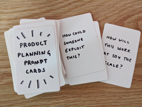 A photo of a stack of blank playing cards. The card at the top of the stack has the words 'Product Planning Prompt Cards' written by hand. A second card says 'How could someone exploit this?' and a third says 'How will this work at 10 times the scale?'