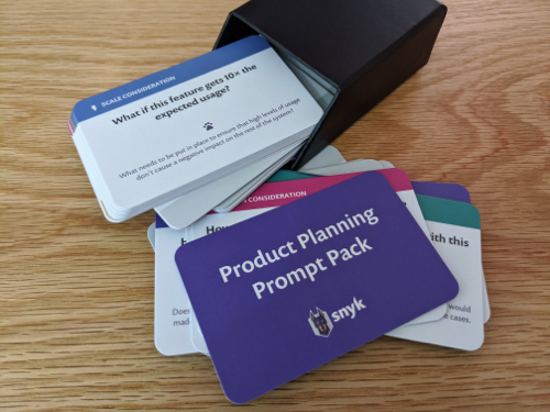 A photo of a stack of printed cards. The card at the top of the stack is purple and has the words 'Product Planning Prompt Pack' printed on it in white text. A second card says 'What if this feature gets 10 times the expected usage?' with smaller text underneath saying 'What needs to be put in place to ensure that high levels of usage don't cause a negative impact on the rest of the system?'